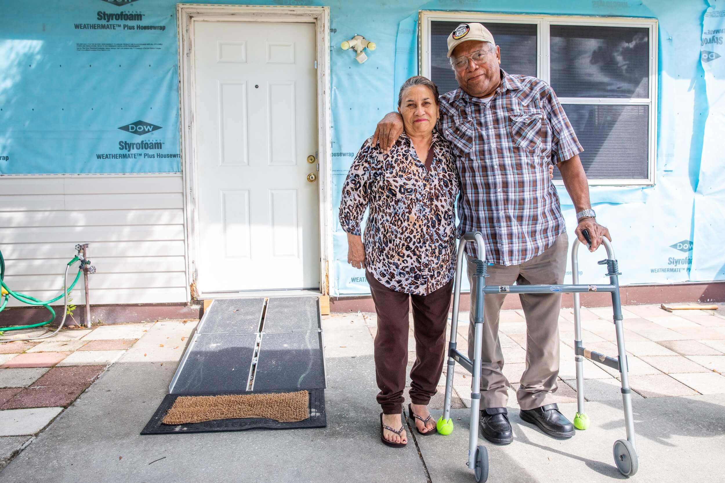 Two older individuals standing in front of a front door, smiling.